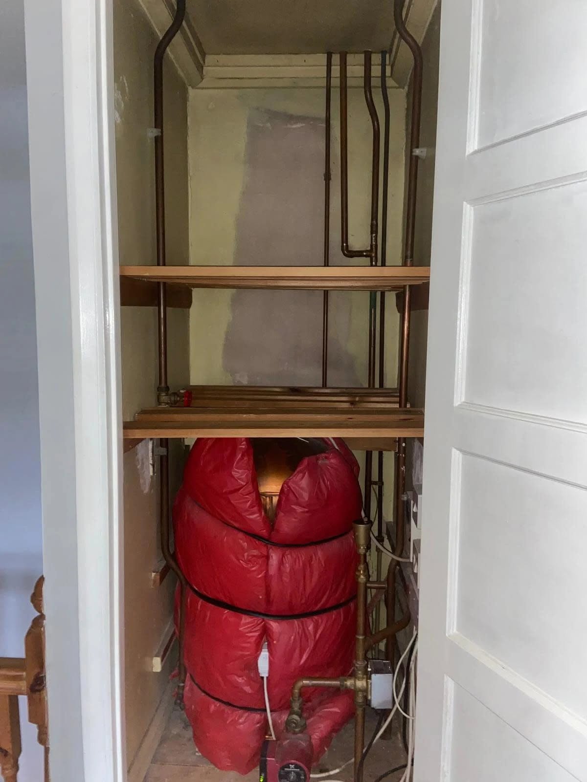An old hot water cylinder in a red insulation jacket inside a cupboard