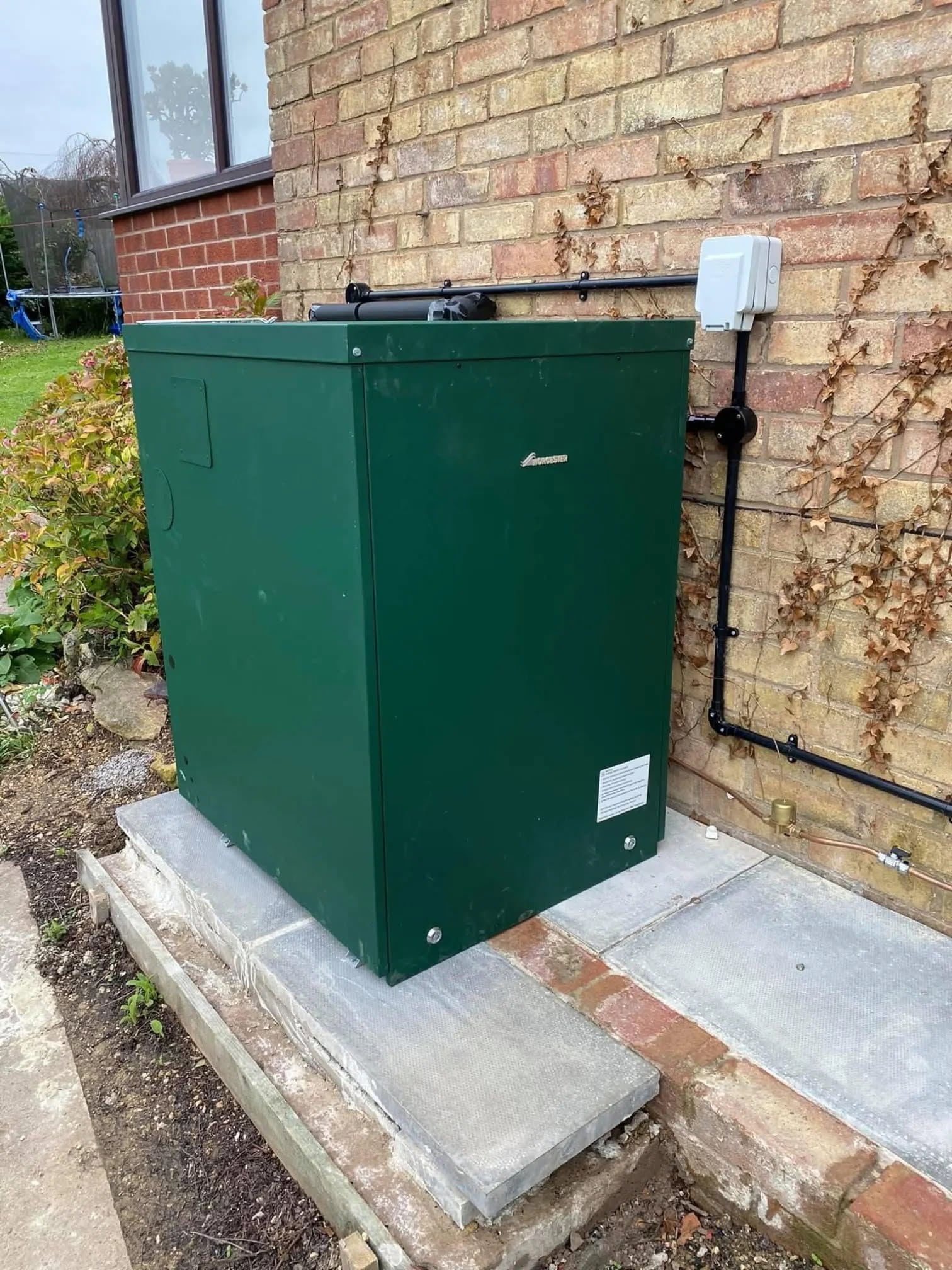 A green external oil boiler on a concrete slab against a brick wall