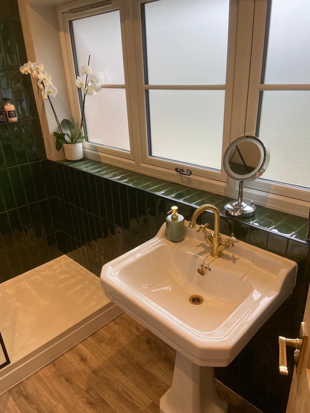 A pedestal basin with brass taps under a window, with green tiles behind